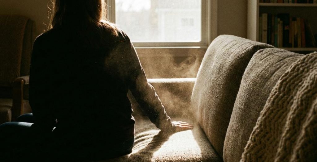 Invisible Allergens: Dust Rising from Furniture Contact Alt Text: A side view of a person sitting down on a textured sofa. Backlighting from a window reveals a subtle cloud of microscopic dust and particulate matter rising into the air from the upholstery upon impact.